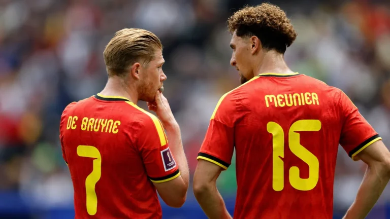 Twee spelers van de Belgische nationale ploeg overleggen op het veld in rode tenues.