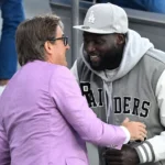 Twee mannen in vrijetijdskleding begroeten elkaar op de tribune van een voetbalstadion.