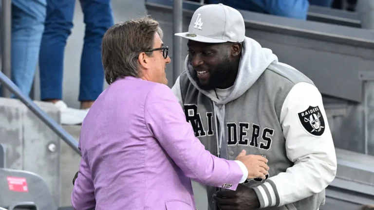 Twee mannen in vrijetijdskleding begroeten elkaar op de tribune van een voetbalstadion.