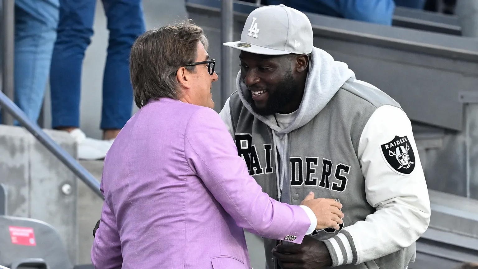 Twee mannen in vrijetijdskleding begroeten elkaar op de tribune van een voetbalstadion.