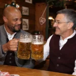 Twee bezoekers in traditionele Beierse kleding heffen bierpullen in een Oktoberfestzaal.