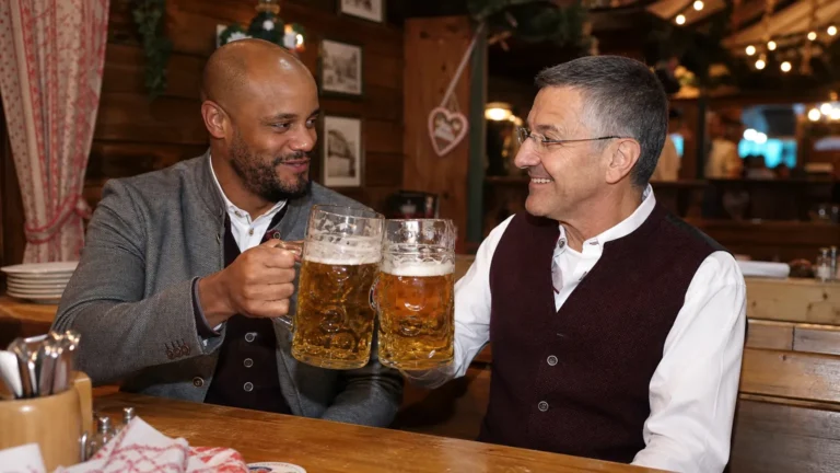 Twee bezoekers in traditionele Beierse kleding heffen bierpullen in een Oktoberfestzaal.