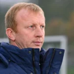 Sven Vandenbroeck in een donkerblauwe jas naast een voetbalveld.