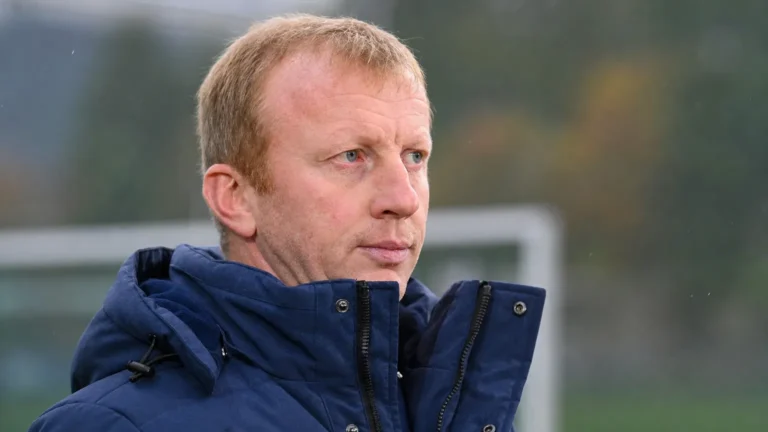 Sven Vandenbroeck in een donkerblauwe jas naast een voetbalveld.