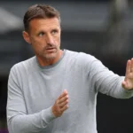 Een OH Leuven-coach in een grijze trui gebaart vanuit de technische zone.