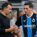 Een Club Brugge-coach overlegt langs het veld met een speler in blauw-zwart tenue.