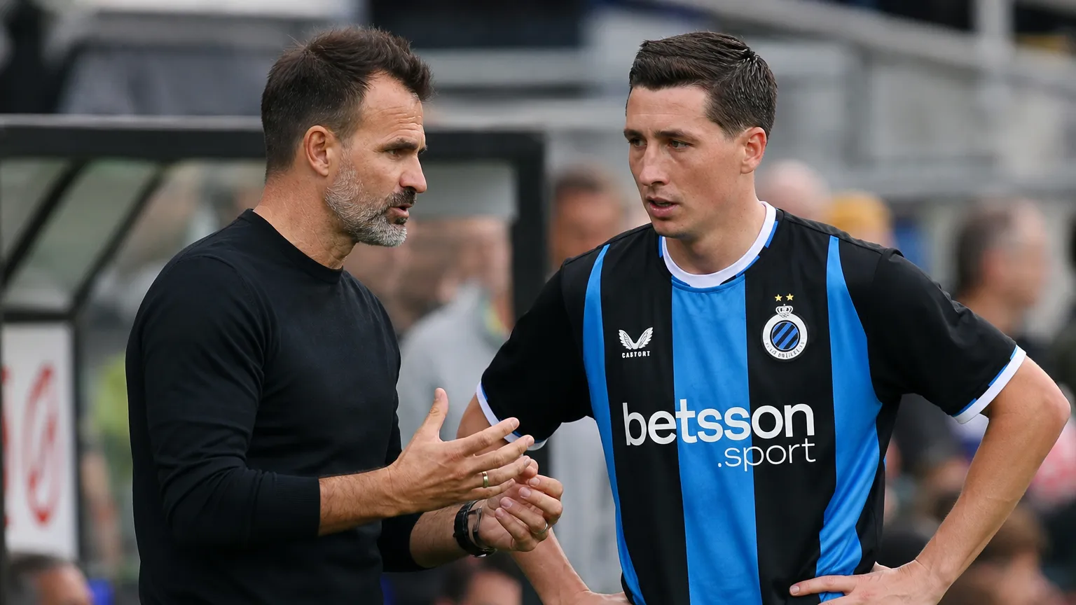Een Club Brugge-coach overlegt langs het veld met een speler in blauw-zwart tenue.