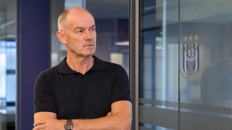 Een niet-spelende clubvertegenwoordiger in een jeugdacademie met een RSCA-embleem op een glazen wand.