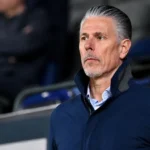 Michael Verschueren kijkt aandachtig toe vanaf de tribune van een voetbalstadion.