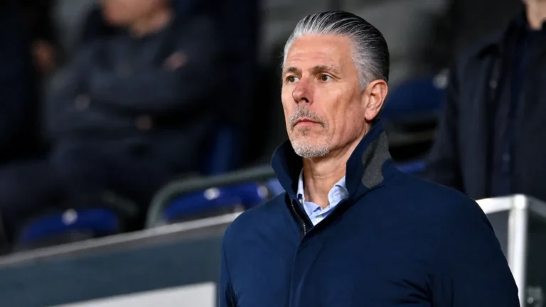 Michael Verschueren kijkt aandachtig toe vanaf de tribune van een voetbalstadion.