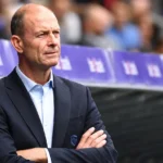 Jess Thorup staat in een donker pak geconcentreerd langs de zijlijn voor een wedstrijd.