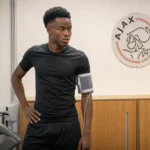 Simon Adingra tijdens een medische controle in een ruimte met een Ajax-wandembleem.