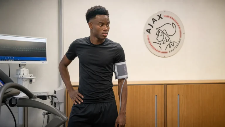 Simon Adingra tijdens een medische controle in een ruimte met een Ajax-wandembleem.