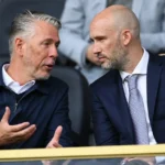 Twee niet-spelende clubvertegenwoordigers overleggen in een tribunezone van een voetbalstadion.
