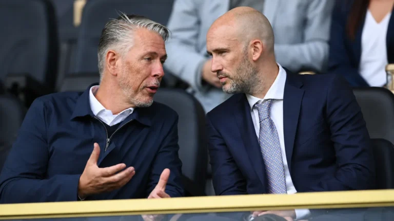 Twee niet-spelende clubvertegenwoordigers overleggen in een tribunezone van een voetbalstadion.