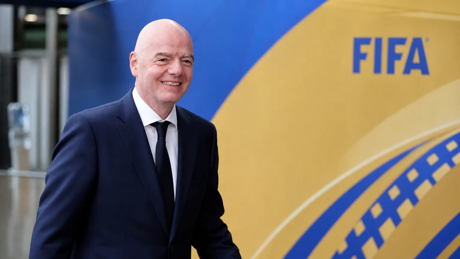 Een FIFA-official in donker pak loopt langs een blauw-gouden FIFA-decor.