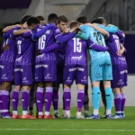 Een groep voetballers in paarse tenues vormt vlak voor de wedstrijd een hechte kring.