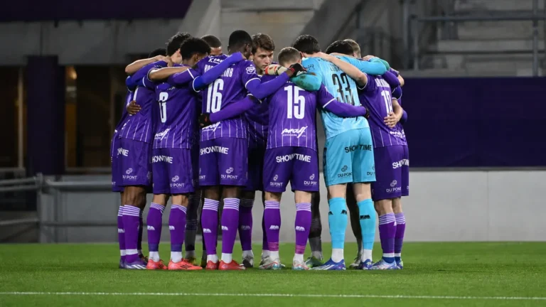 Een groep voetballers in paarse tenues vormt vlak voor de wedstrijd een hechte kring.