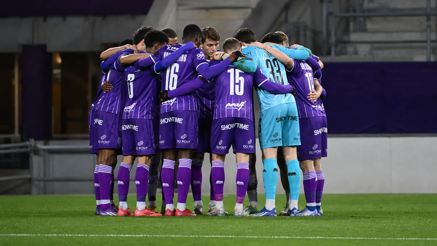 Een groep voetballers in paarse tenues vormt vlak voor de wedstrijd een hechte kring.