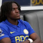 Een speler in blauw shirt zit op de spelersbank van een voetbalstadion.