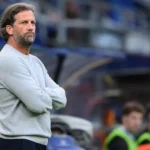 Een KAA Gent-coach in een lichtgrijze trui staat geconcentreerd langs het veld.