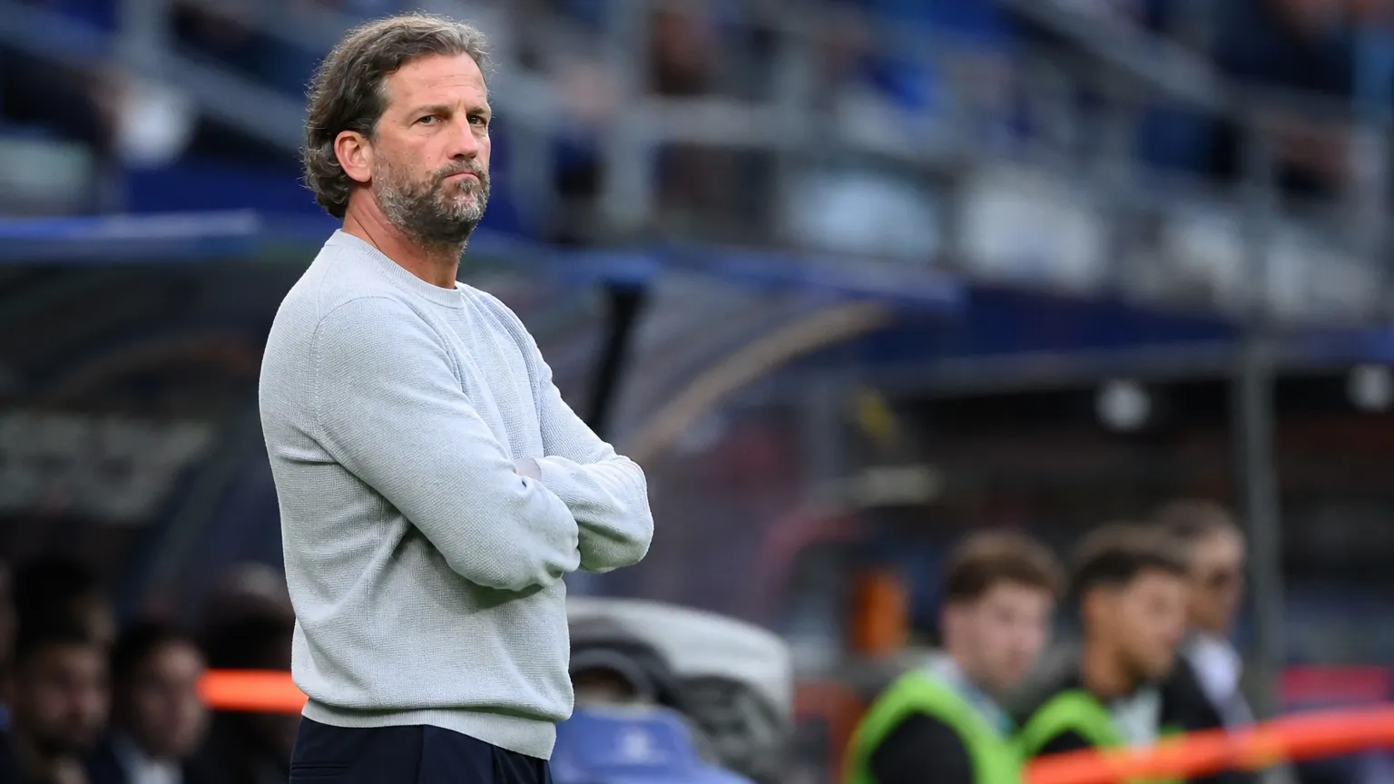 Een KAA Gent-coach in een lichtgrijze trui staat geconcentreerd langs het veld.
