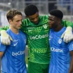 Drie voetballers in blauwe en groene wedstrijdtenues op het veld.