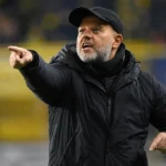Een coach in donkere jas geeft instructies langs de zijlijn van een voetbalstadion.