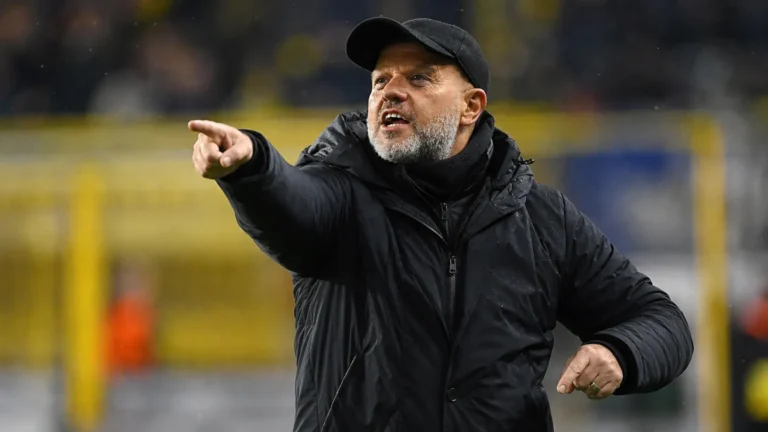Een coach in donkere jas geeft instructies langs de zijlijn van een voetbalstadion.