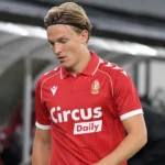 Een voetballer in een rood shirt loopt teleurgesteld langs de zijlijn na een wedstrijd.