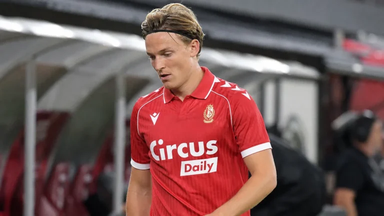 Een voetballer in een rood shirt loopt teleurgesteld langs de zijlijn na een wedstrijd.
