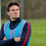 Jelle Vossen kijkt vanaf de zijlijn naar het veld in een donker trainingsvest en blauw hesje.