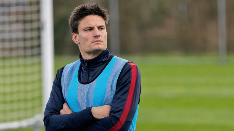 Jelle Vossen kijkt vanaf de zijlijn naar het veld in een donker trainingsvest en blauw hesje.