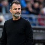 Een coach in zwarte trui kijkt aandachtig toe vanaf de zijlijn van een stadion.