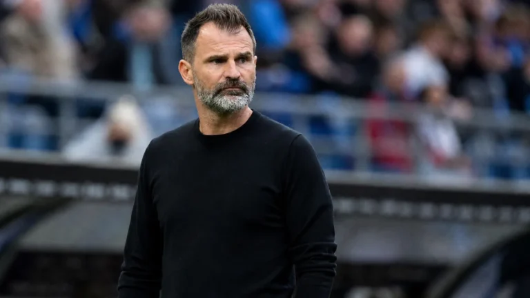 Een coach in zwarte trui kijkt aandachtig toe vanaf de zijlijn van een stadion.