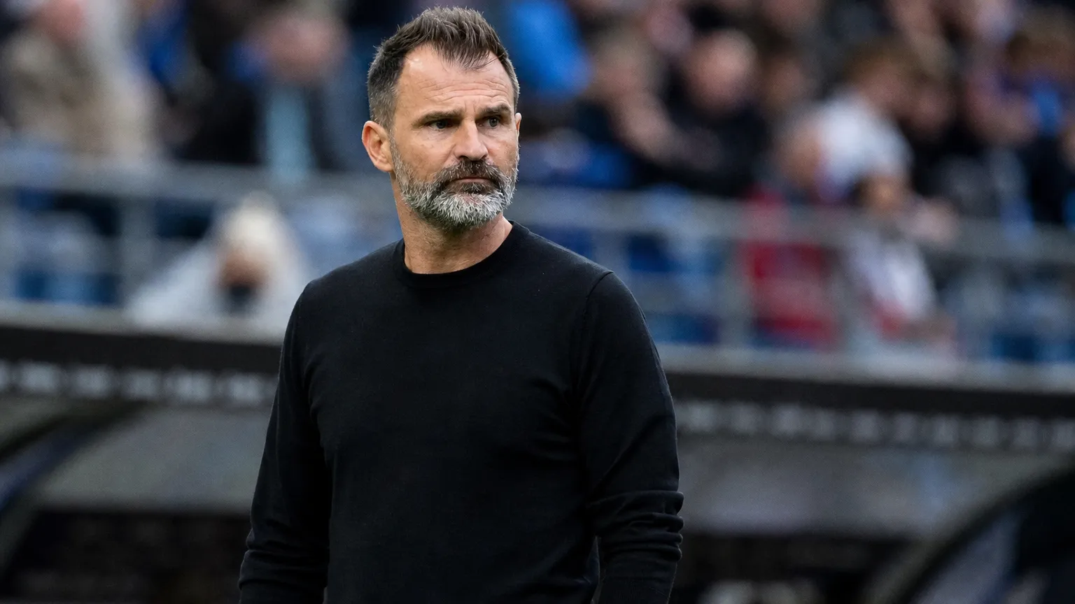 Een coach in zwarte trui kijkt aandachtig toe vanaf de zijlijn van een stadion.