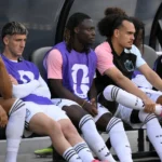 Een groep Belgische wisselspelers zit in witte tenues en paarse hesjes op de bank van een dug-out.