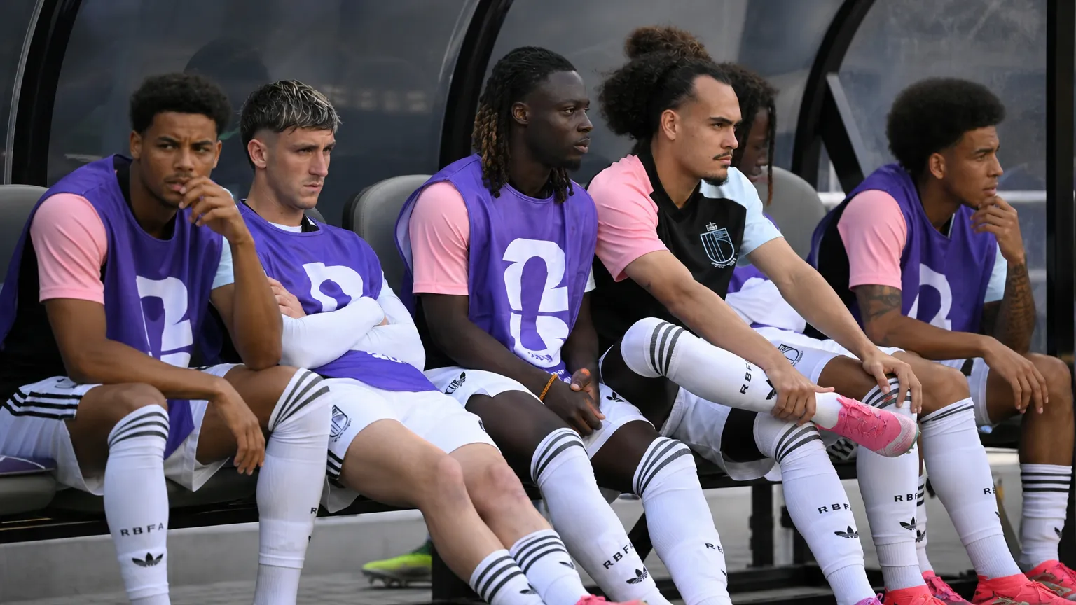 Een groep Belgische wisselspelers zit in witte tenues en paarse hesjes op de bank van een dug-out.