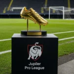 Een gouden topschutterstrofee op een plint naast een voetbalveld met een Jupiler Pro League-embleem.