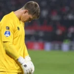 Een doelman in geel staat met gebogen hoofd op het veld tijdens een steunbetuiging.