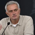 Een trainer in beige polo spreekt na een wedstrijd op een persconferentie.