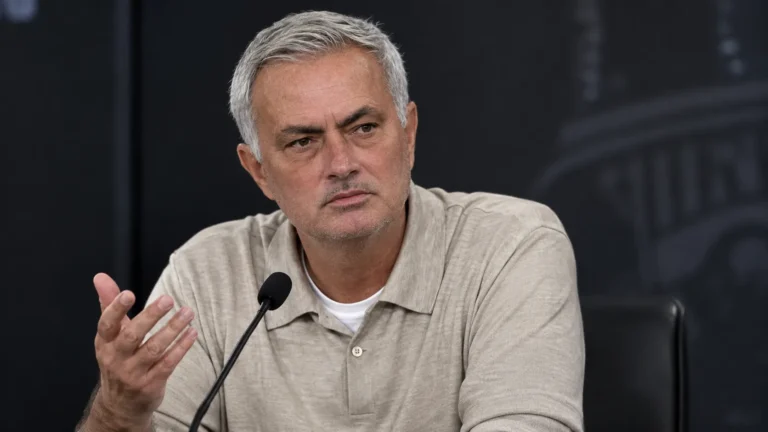 Een trainer in beige polo spreekt na een wedstrijd op een persconferentie.
