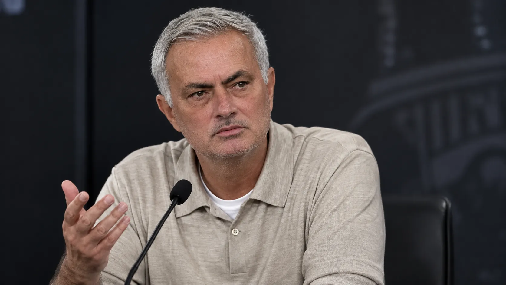 Een trainer in beige polo spreekt na een wedstrijd op een persconferentie.