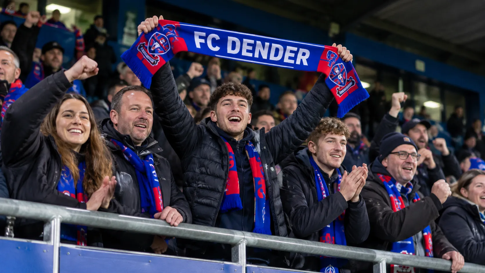 Een collectieve groep FC Dender-supporters viert in de tribune met een blauw-rode sjaal.
