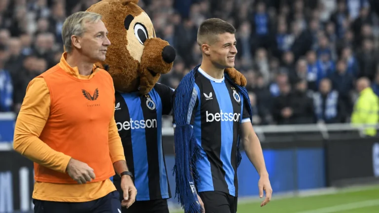 Een speler in blauw-zwart tenue loopt na de wedstrijd langs de zijlijn met een mascotte en een staflid.