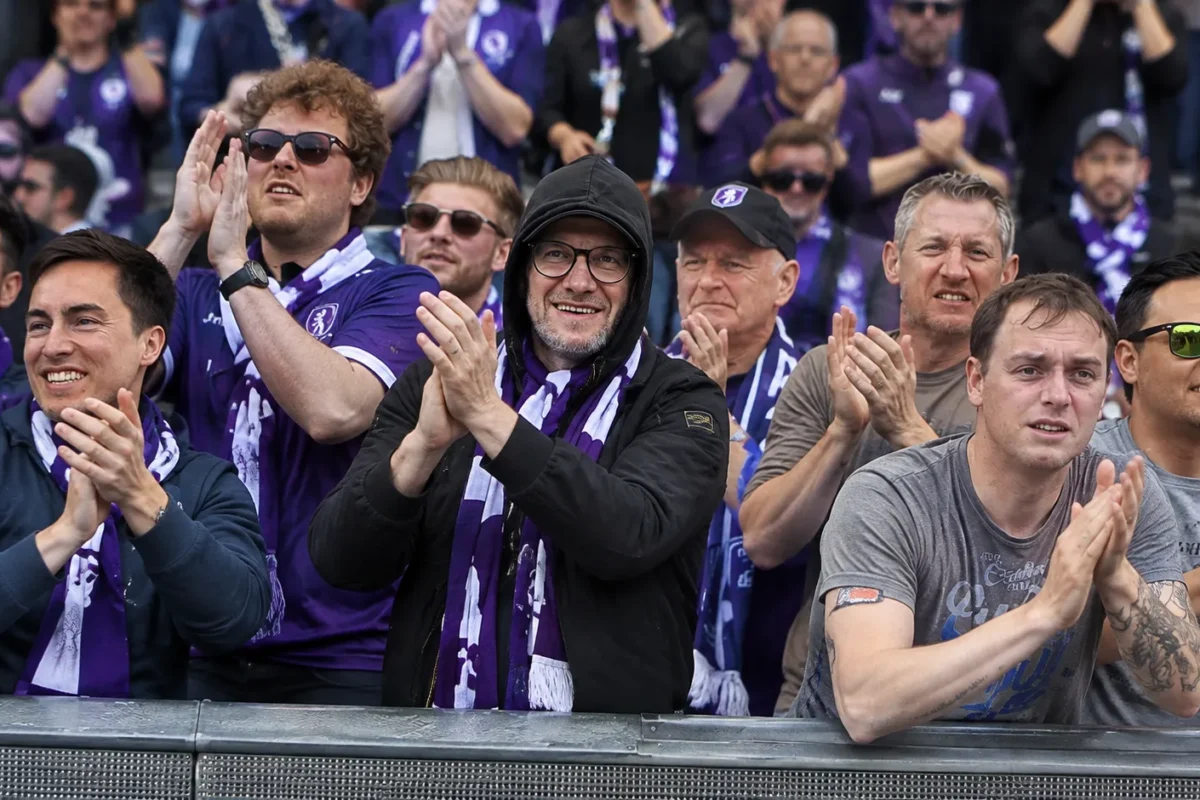 Een groep supporters in paarse voetbalshirts applaudisseert achter een stadionafsluiting.