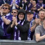 Een groep supporters in paarse voetbalshirts applaudisseert achter een stadionafsluiting.