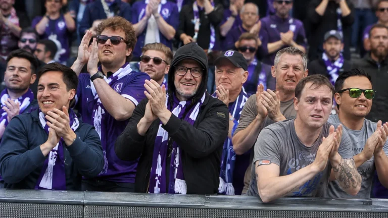 Een groep supporters in paarse voetbalshirts applaudisseert achter een stadionafsluiting.