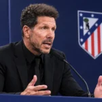 Diego Simeone in zwart pak tijdens een persconferentie, met een Atlético Madrid-embleem op de achterwand.