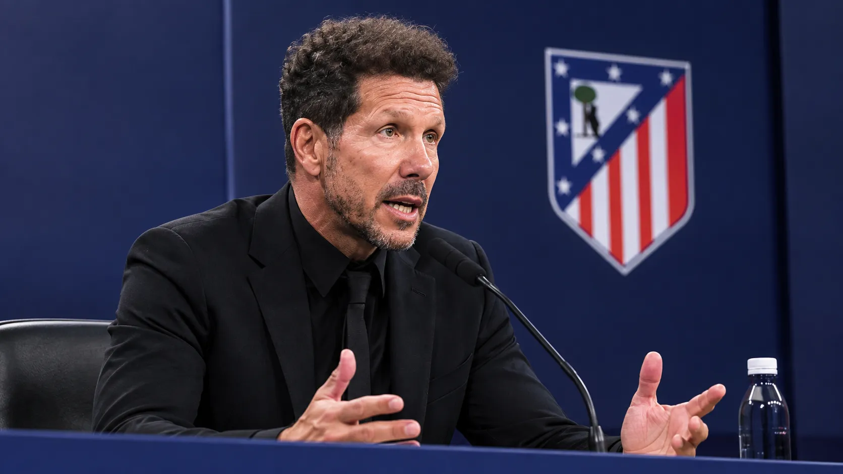 Diego Simeone in zwart pak tijdens een persconferentie, met een Atlético Madrid-embleem op de achterwand.